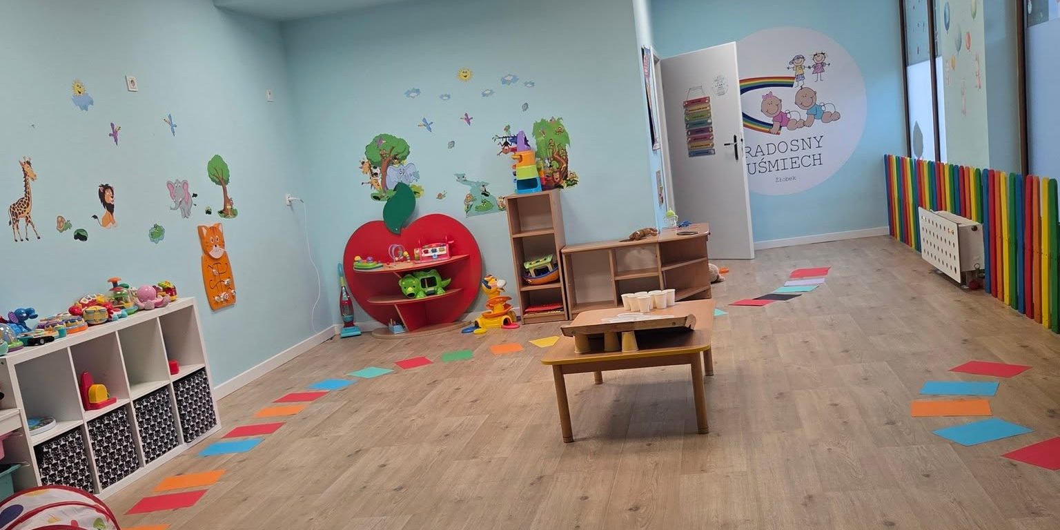Sala zabaw żłobka Radosny Uśmiech z tęczowym płotkiem, kolorowymi zabawkami i logo na ścianie